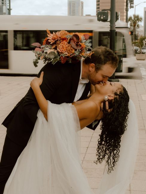 A romantic wedding moment featuring a groom in a black suit holding a bride in a white wedding gown and veil, as they share a dance or intimate embrace outdoors in front of a busy urban street scene.